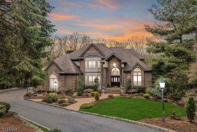 french provincial home with brick siding, a shingled roof, driveway, a lawn, and a chimney