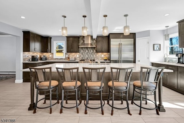 kitchen featuring tasteful backsplash, a baseboard radiator, stainless steel built in refrigerator, dark brown cabinets, and wall chimney range hood