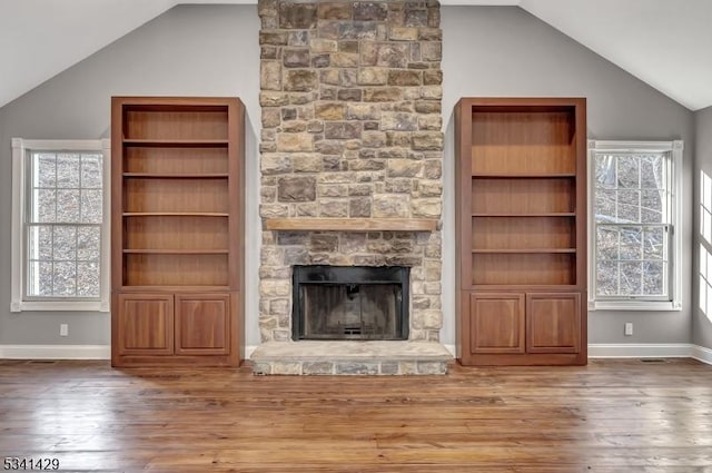 unfurnished living room featuring a stone fireplace, built in shelves, wood finished floors, baseboards, and vaulted ceiling