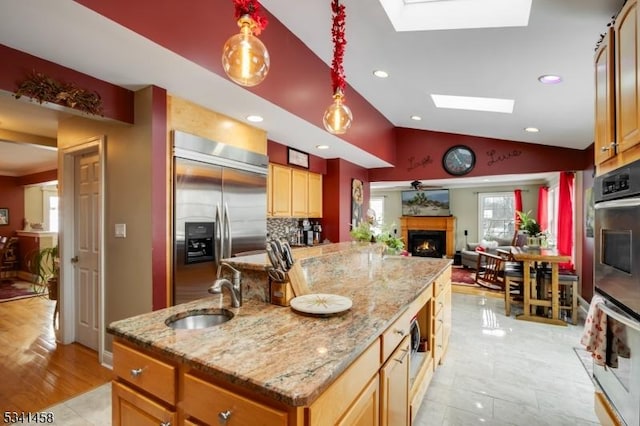 kitchen with vaulted ceiling with skylight, a center island with sink, a lit fireplace, stainless steel built in refrigerator, and a sink