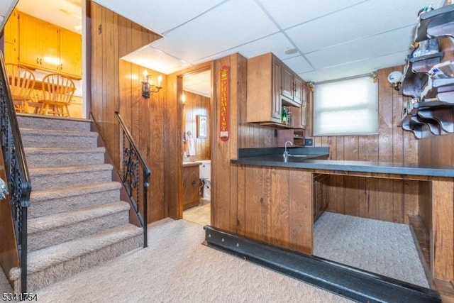 kitchen with wooden walls, brown cabinetry, carpet, a paneled ceiling, and a sink