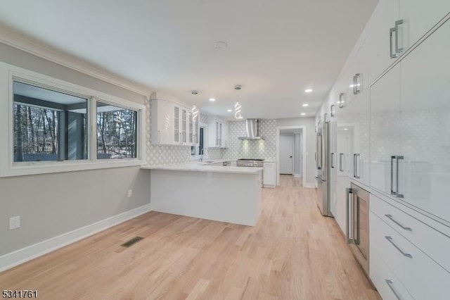 kitchen featuring white cabinets, glass insert cabinets, freestanding refrigerator, a peninsula, and wall chimney range hood