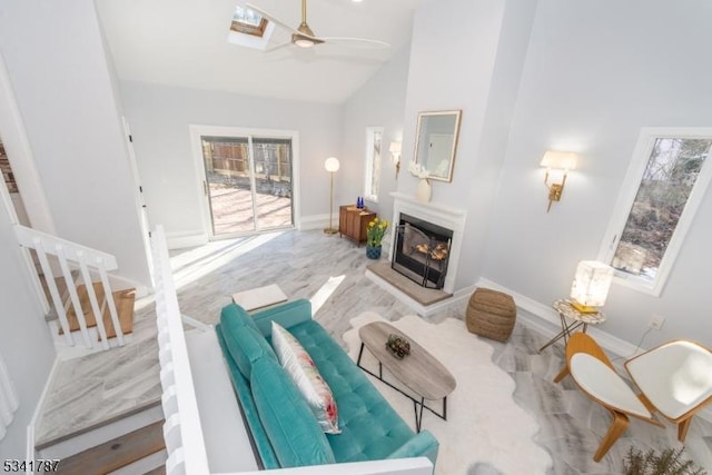 living area with high vaulted ceiling, a skylight, a ceiling fan, baseboards, and a glass covered fireplace