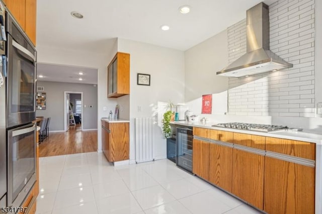 kitchen with beverage cooler, brown cabinetry, radiator, wall chimney exhaust hood, and stainless steel double oven