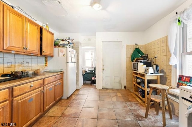 kitchen featuring tasteful backsplash, stainless steel microwave, brown cabinetry, freestanding refrigerator, and light tile patterned flooring