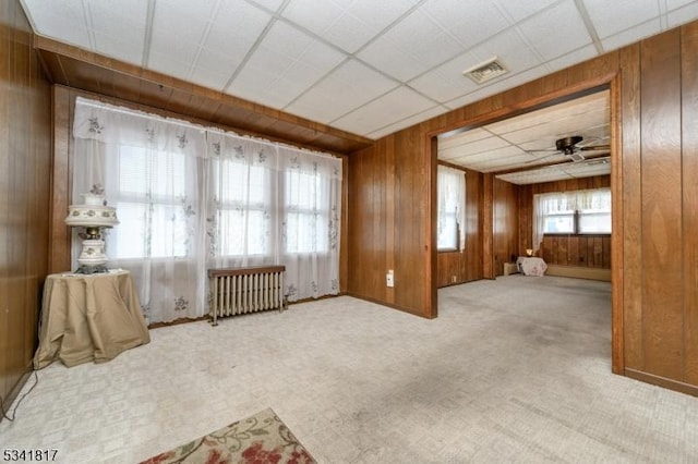 carpeted empty room with ceiling fan, a drop ceiling, wood walls, visible vents, and radiator heating unit