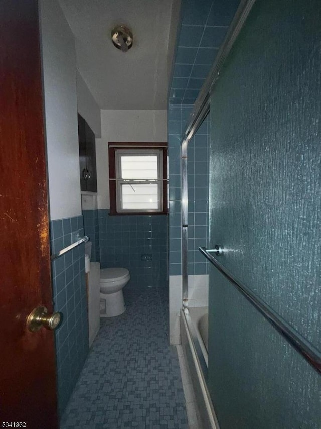 full bath with a wainscoted wall, tile walls, toilet, and bath / shower combo with glass door