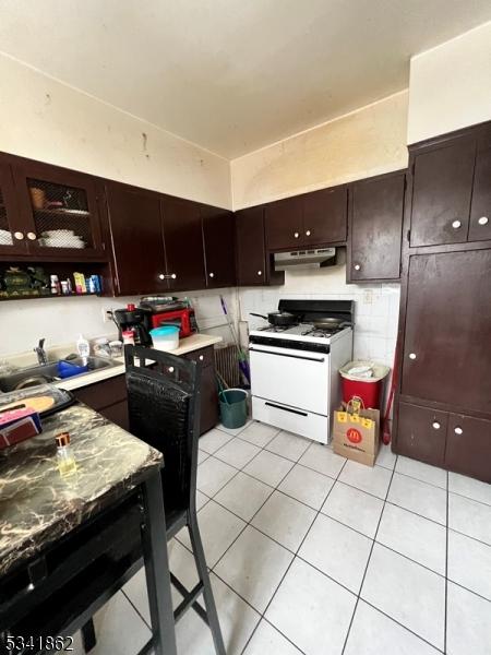 kitchen featuring light tile patterned floors, white range with gas stovetop, dark brown cabinets, under cabinet range hood, and a sink