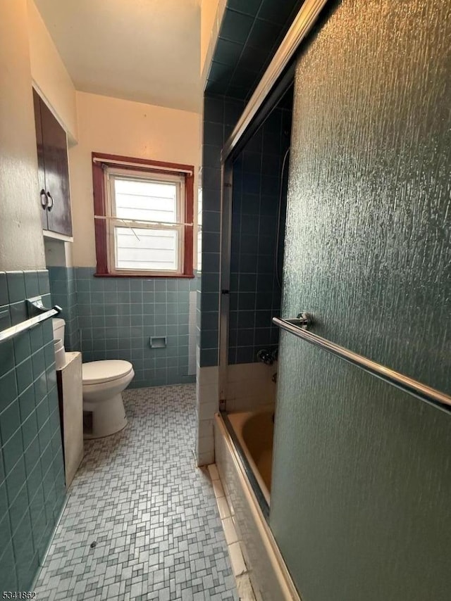 full bath with wainscoting, toilet, tile patterned flooring, combined bath / shower with glass door, and tile walls