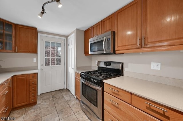 kitchen featuring brown cabinets, light countertops, appliances with stainless steel finishes, glass insert cabinets, and light tile patterned flooring