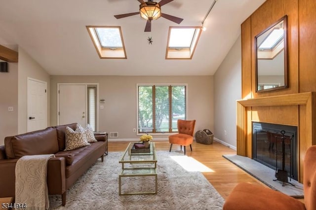 living area with a ceiling fan, visible vents, light wood finished floors, vaulted ceiling with skylight, and a glass covered fireplace