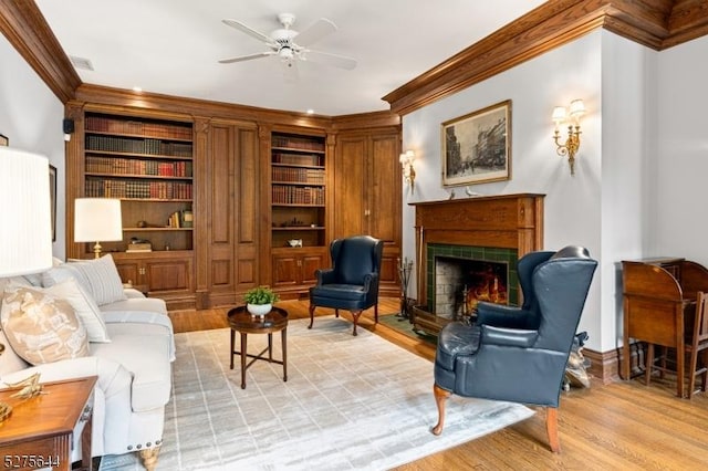 living area featuring ceiling fan, a tile fireplace, built in features, light wood finished floors, and crown molding