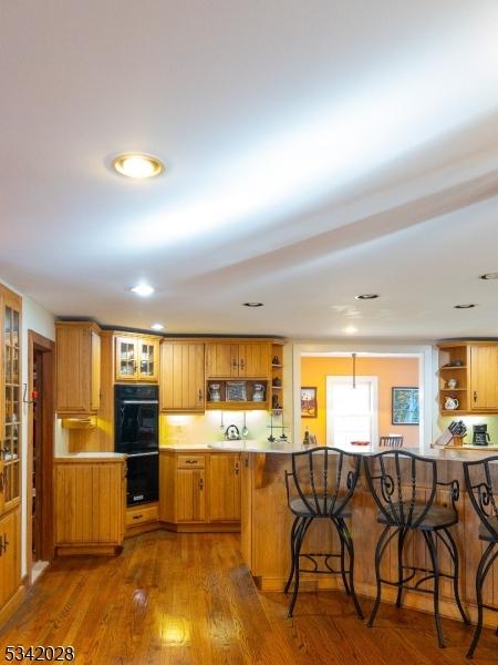 kitchen with brown cabinets, dark wood-type flooring, dobule oven black, open shelves, and a kitchen breakfast bar