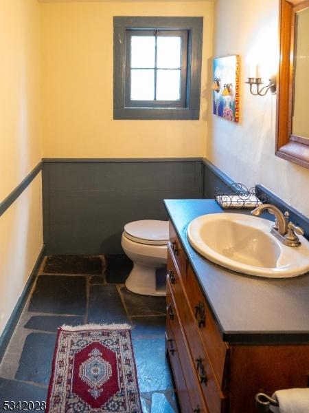 half bathroom featuring a wainscoted wall, toilet, vanity, and stone finish floor