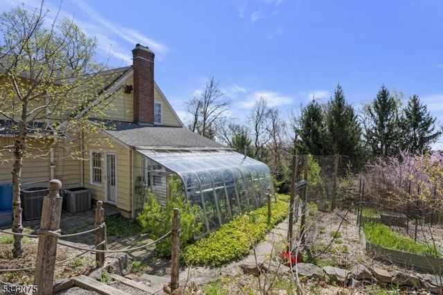 view of side of property with a chimney, central AC unit, roof with shingles, and a greenhouse