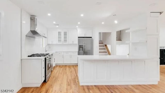 kitchen featuring wall chimney exhaust hood, light wood-style flooring, appliances with stainless steel finishes, light countertops, and white cabinetry