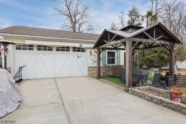 exterior space with a garage, a shingled roof, concrete driveway, stone siding, and a gazebo