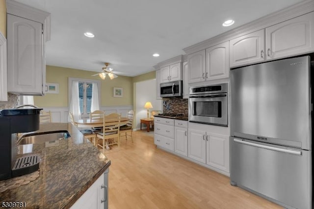 kitchen with dark stone counters, wainscoting, light wood-style flooring, appliances with stainless steel finishes, and a ceiling fan