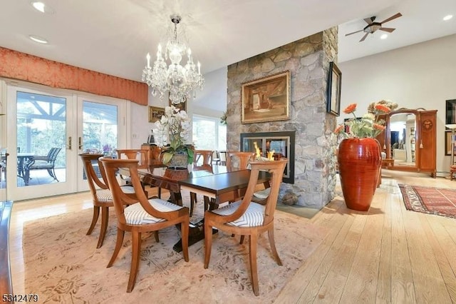 dining space featuring light wood-style flooring, ceiling fan with notable chandelier, french doors, and a stone fireplace