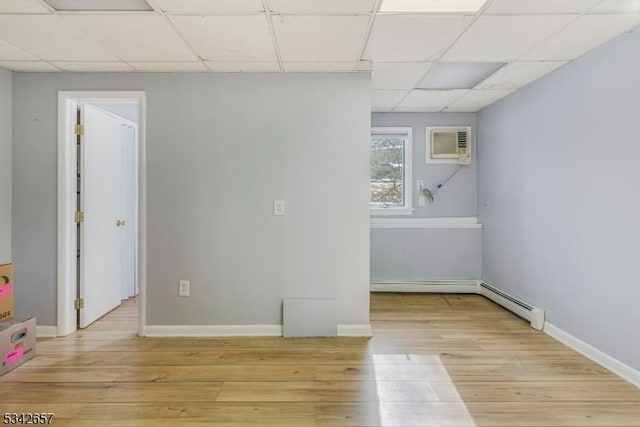 empty room with light wood-style flooring, baseboard heating, an AC wall unit, a drop ceiling, and baseboards