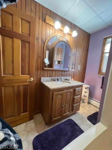 bathroom featuring baseboard heating, a drop ceiling, vanity, and wooden walls