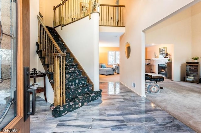 carpeted entrance foyer featuring a warm lit fireplace, stairs, a towering ceiling, and baseboards