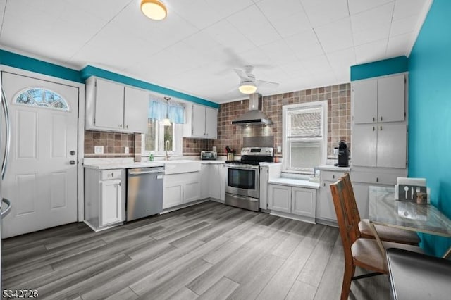 kitchen featuring light countertops, decorative backsplash, appliances with stainless steel finishes, a sink, and wall chimney exhaust hood