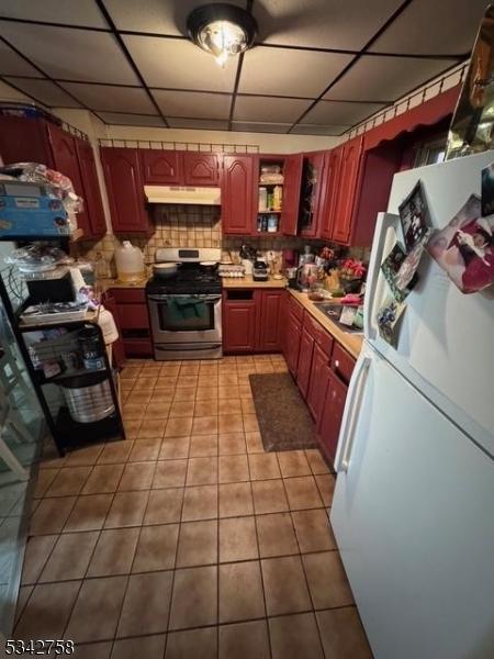 kitchen featuring stainless steel gas stove, light tile patterned floors, freestanding refrigerator, a paneled ceiling, and under cabinet range hood