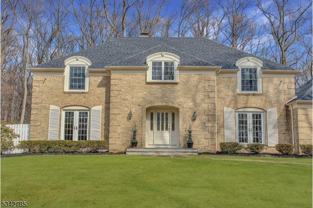 french country style house with a front lawn, french doors, roof with shingles, brick siding, and a chimney