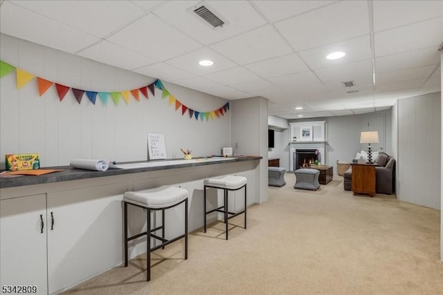 kitchen with visible vents, a drop ceiling, light colored carpet, a kitchen bar, and a warm lit fireplace