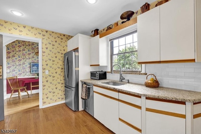 kitchen with appliances with stainless steel finishes, a sink, white cabinets, light wood-style floors, and wallpapered walls