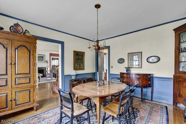dining area with wood finished floors, a wainscoted wall, ornamental molding, a brick fireplace, and a chandelier
