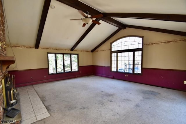 unfurnished room with ceiling fan, lofted ceiling with beams, carpet flooring, wainscoting, and a fireplace