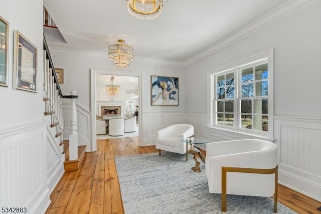 sitting room featuring stairway, a wainscoted wall, light wood-style flooring, a warm lit fireplace, and crown molding