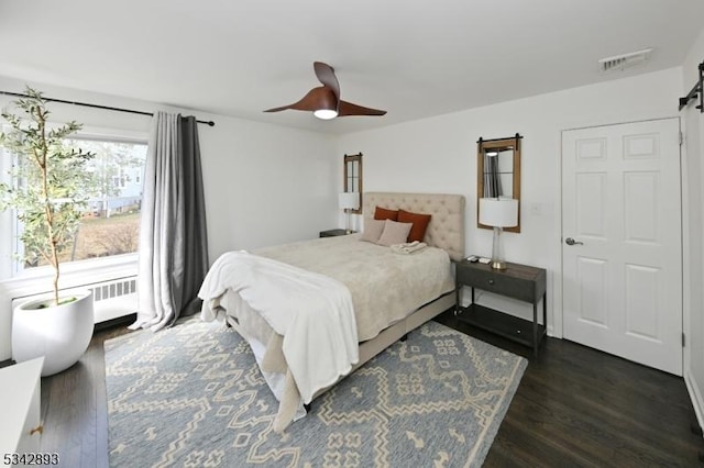 bedroom featuring visible vents, ceiling fan, radiator heating unit, a barn door, and dark wood-style flooring