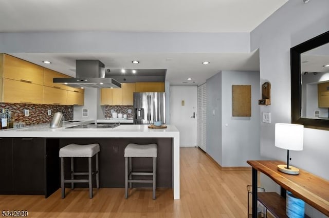 kitchen with a peninsula, light countertops, stainless steel refrigerator with ice dispenser, tasteful backsplash, and island range hood