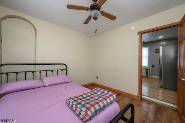 bedroom featuring radiator, baseboards, freestanding refrigerator, wood finished floors, and a ceiling fan