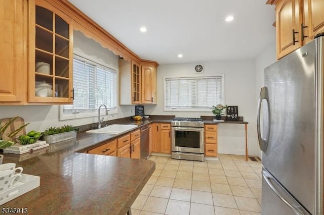 kitchen with a sink, dark countertops, appliances with stainless steel finishes, light tile patterned floors, and glass insert cabinets