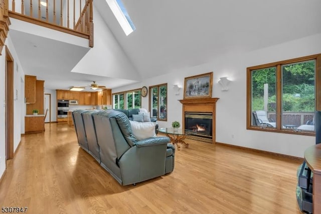 living room with baseboards, light wood-type flooring, a skylight, a glass covered fireplace, and high vaulted ceiling