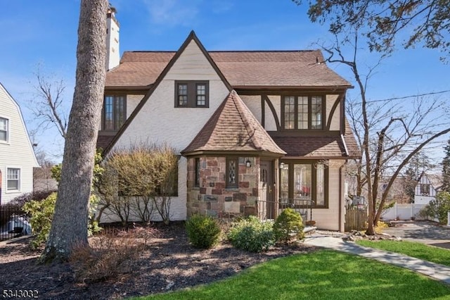 english style home with stone siding, a shingled roof, and fence