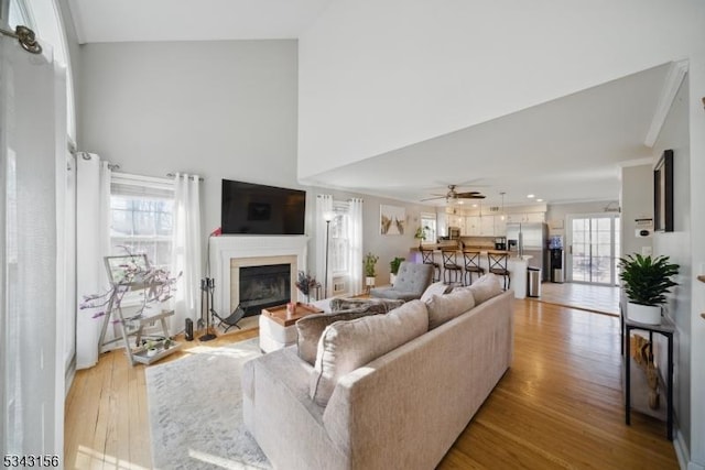 living area with a glass covered fireplace, light wood-style floors, a towering ceiling, and ceiling fan