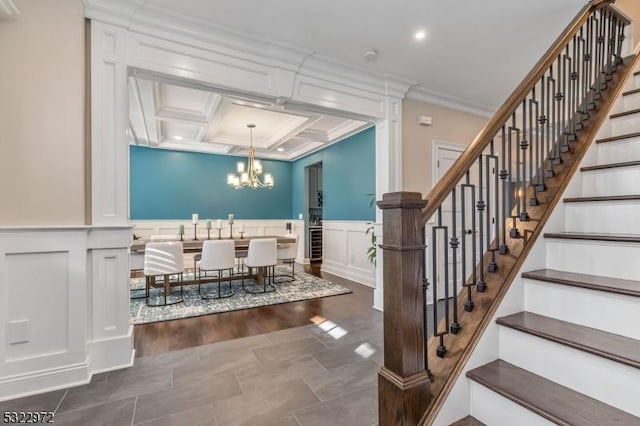 staircase featuring coffered ceiling, wainscoting, crown molding, a decorative wall, and a notable chandelier