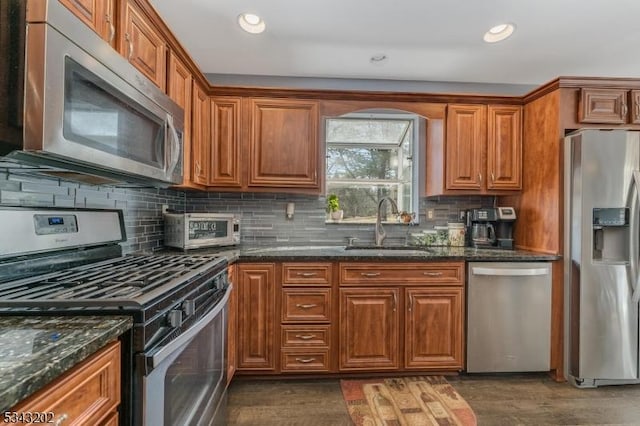 kitchen featuring brown cabinetry, dark stone counters, a sink, stainless steel appliances, and backsplash