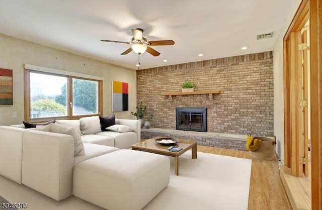 living room featuring visible vents, light wood-style flooring, brick wall, a brick fireplace, and ceiling fan