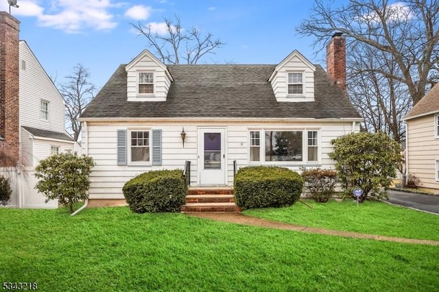 new england style home with entry steps, a chimney, a front lawn, and a shingled roof