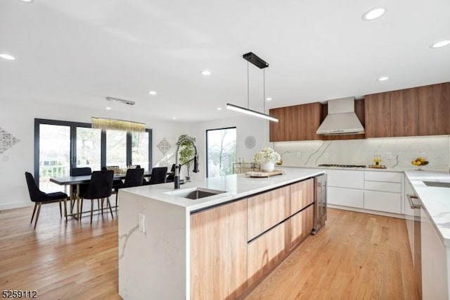 kitchen featuring modern cabinets, an island with sink, a sink, wall chimney range hood, and a healthy amount of sunlight