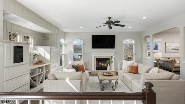 living room with a ceiling fan, recessed lighting, arched walkways, and a warm lit fireplace