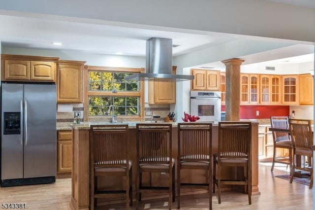 kitchen with ornate columns, stainless steel appliances, glass insert cabinets, light wood-type flooring, and island range hood