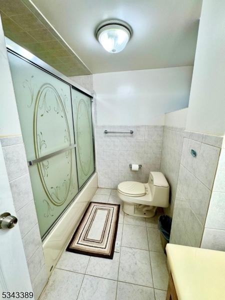 full bath with a wainscoted wall, shower / bath combination with glass door, tile patterned floors, toilet, and tile walls