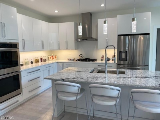 kitchen with a breakfast bar, a sink, appliances with stainless steel finishes, wall chimney range hood, and hanging light fixtures